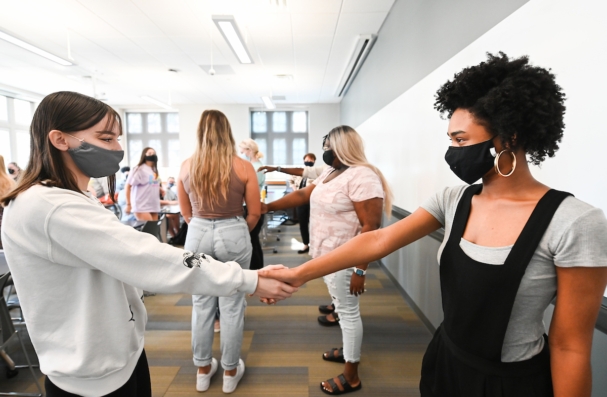 Two women in a classroom, wearing face masks, hold hands. Others in the background do the same.