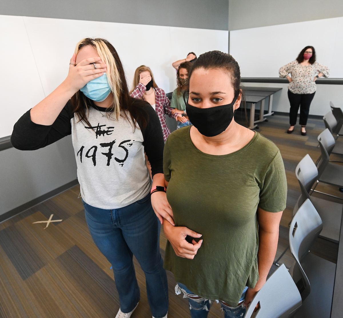 A woman in a green shirt and black mask leads another with a "The 1975" shirt, eyes covered, in a classroom. Others follow in similar pairs.