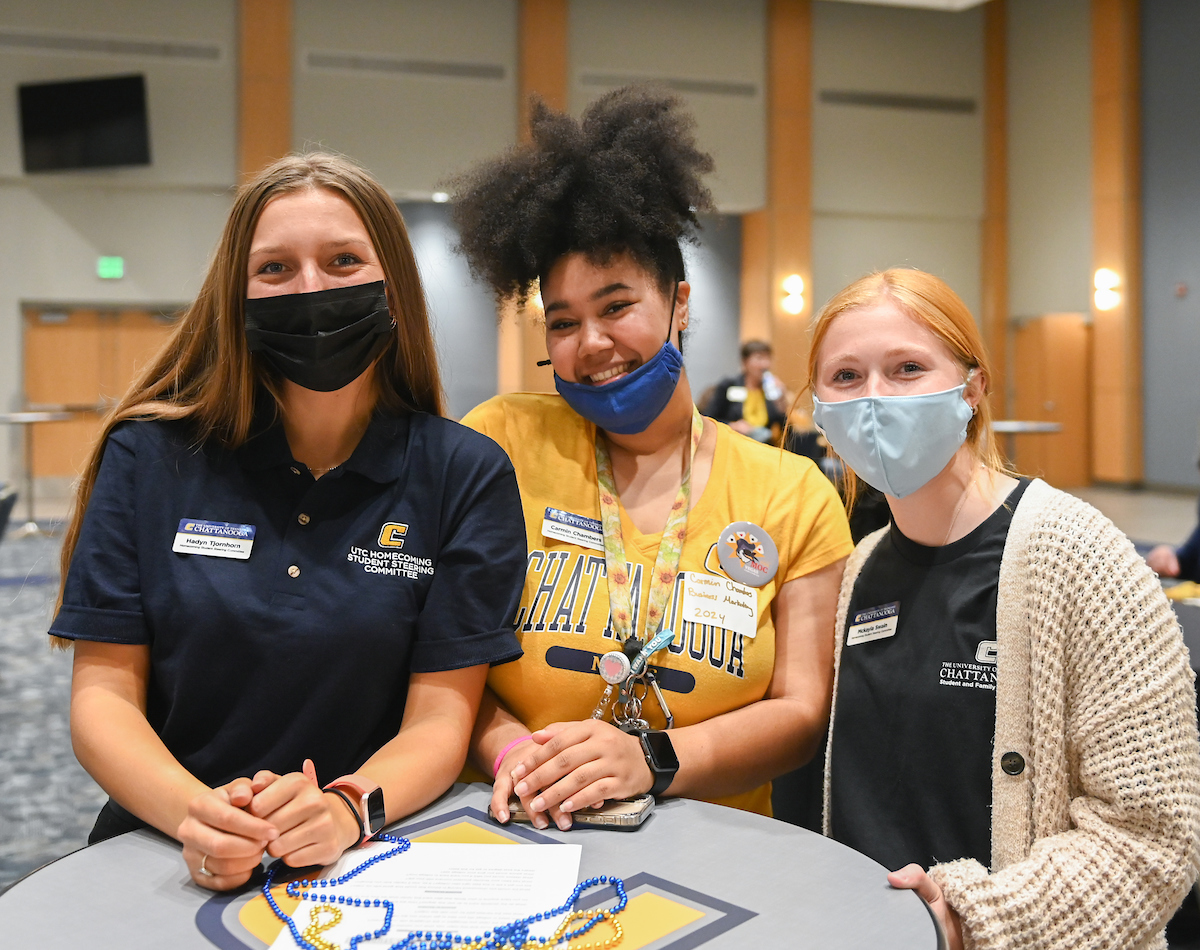 Three young women wearing masks and casual attire smile at a table with beads in a bright indoor setting.