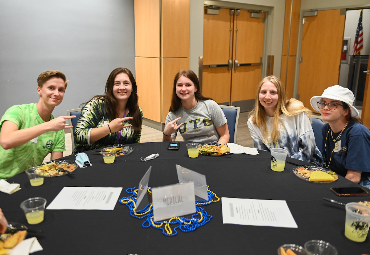 A group of students sit around a table with plates of food and drinks, smiling and holding up the "Power C".