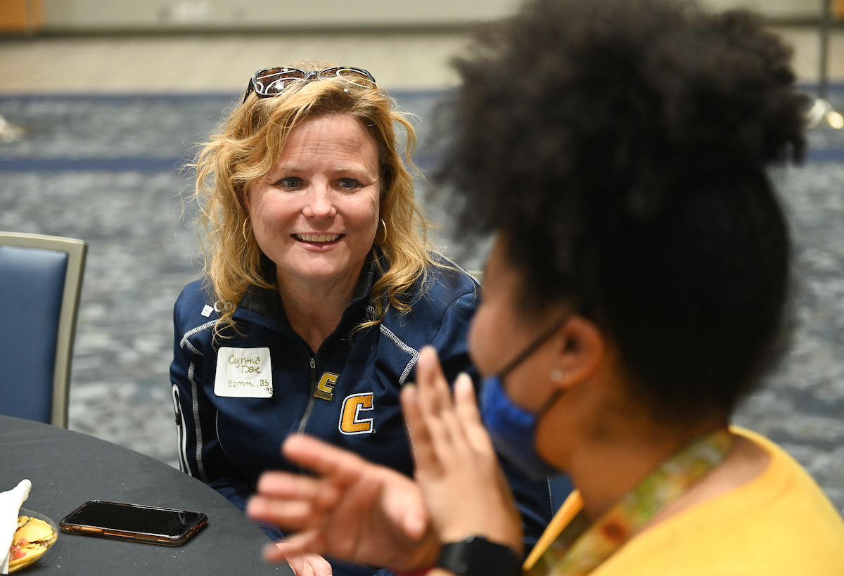 A woman sits at a table, smiling while engaging in conversation with another woman wearing a mask.