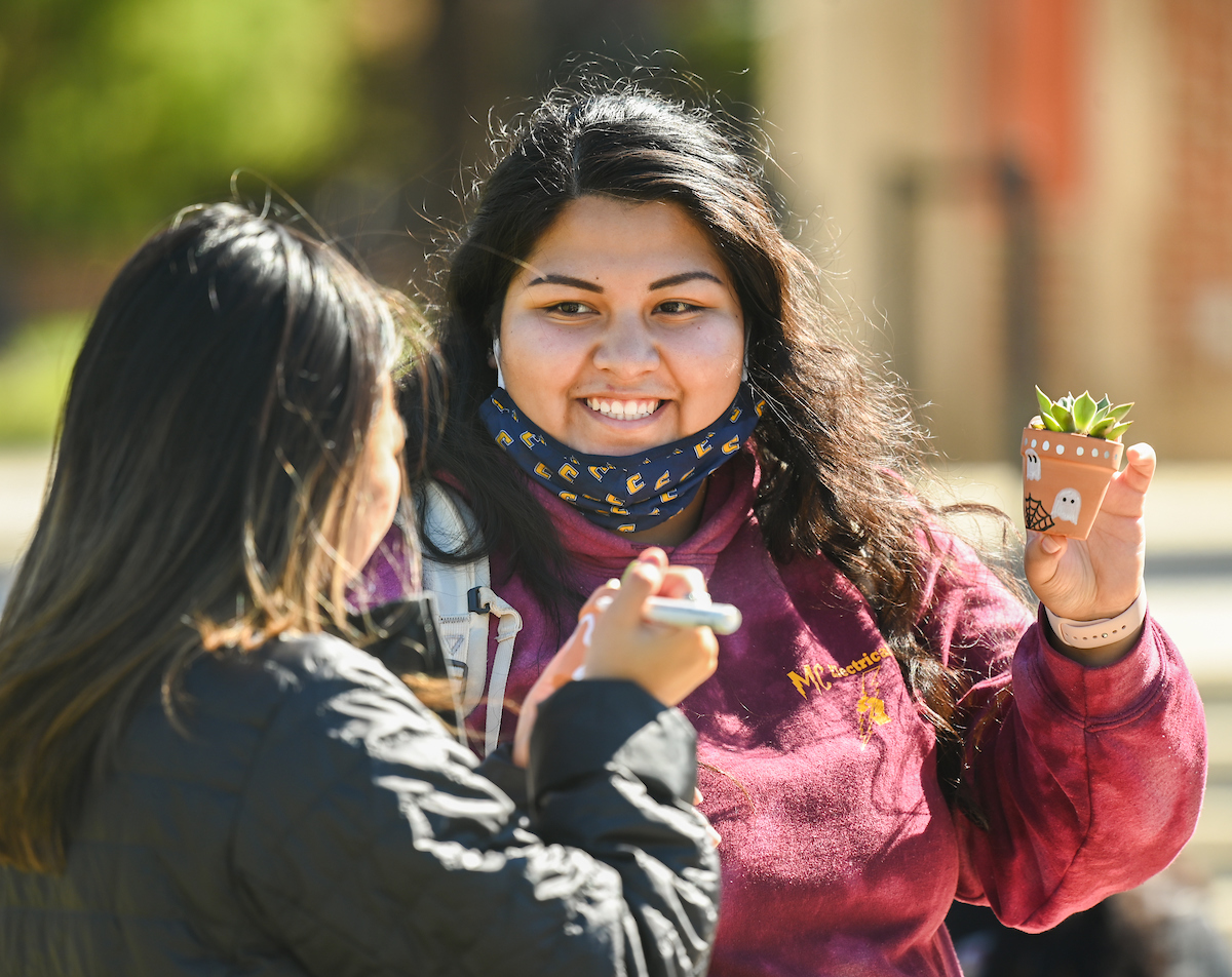 Two students outdoors, smiling. One holds a small potted plant decorated with a ghost.