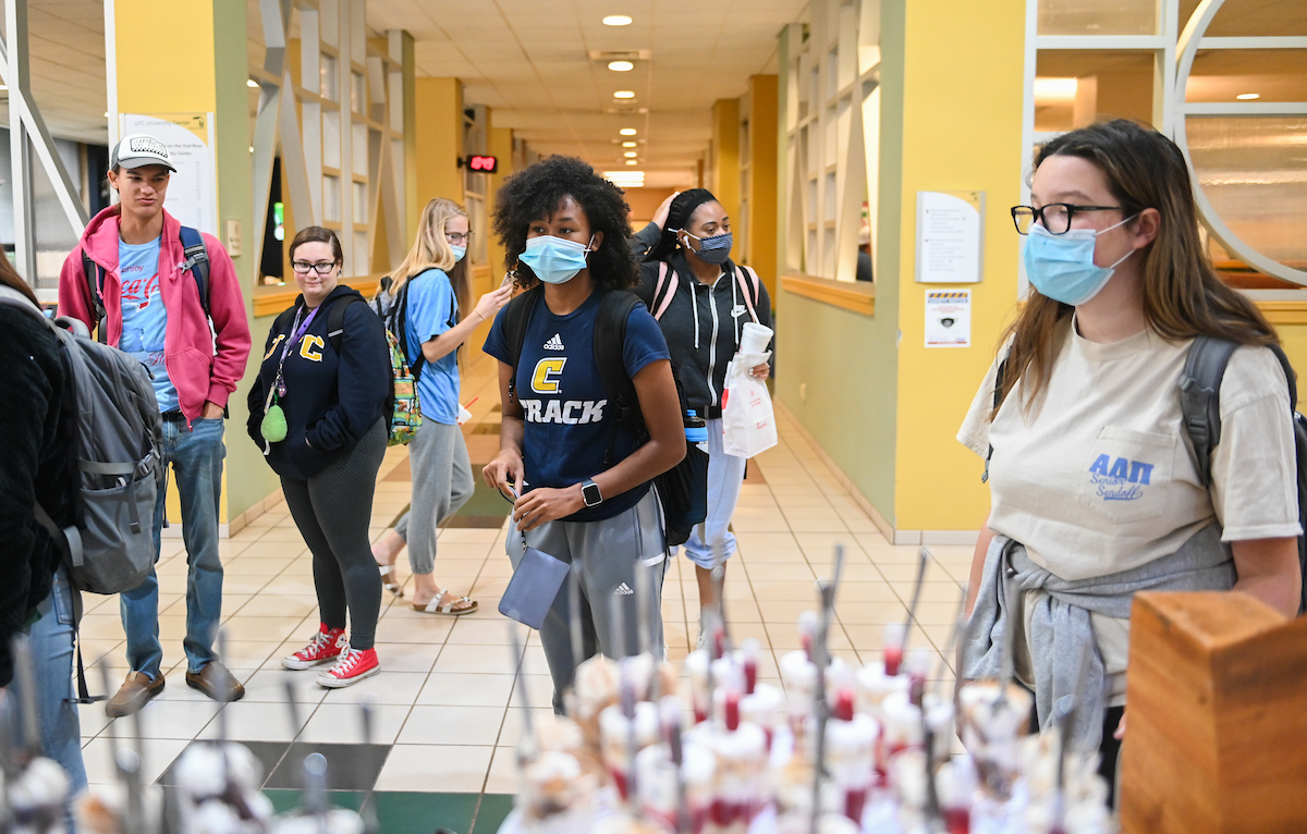 Students wearing masks stand in a bright hallway, carrying backpacks. With a display of snacks in the foreground.