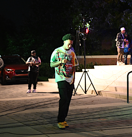 A person in a colorful shirt holds a microphone, standing near steps under a spotlight at night. A small group gathers in the background.