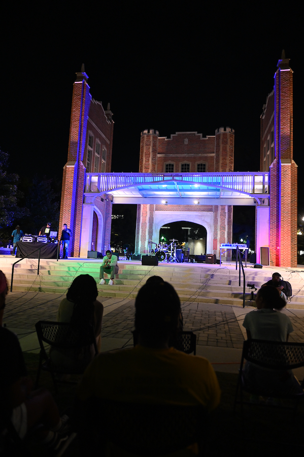Outdoor nighttime concert with colorful lights illuminating a brick stage. A performer kneels and sings, while an audience watches.