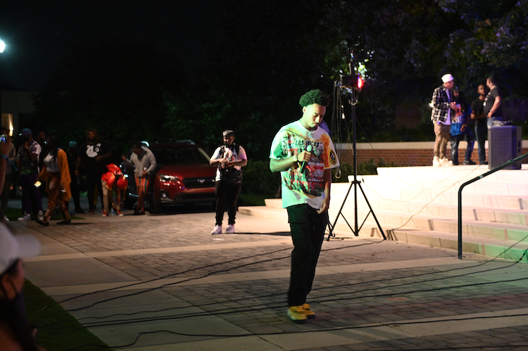 A person in a colorful shirt holds a microphone, standing near steps under a spotlight at night. A small group gathers in the background.