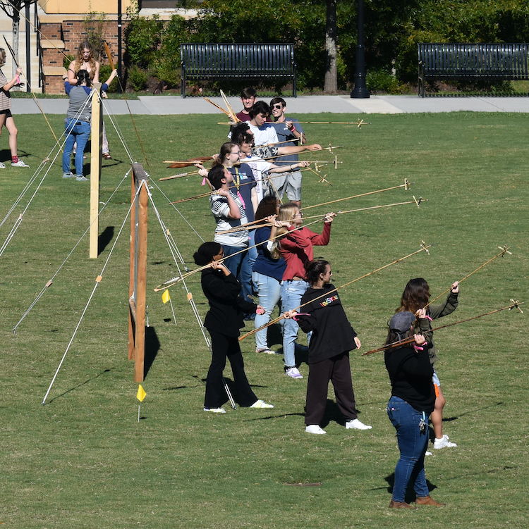 A group of students throwing atlatls into the sky in a grassy field.
