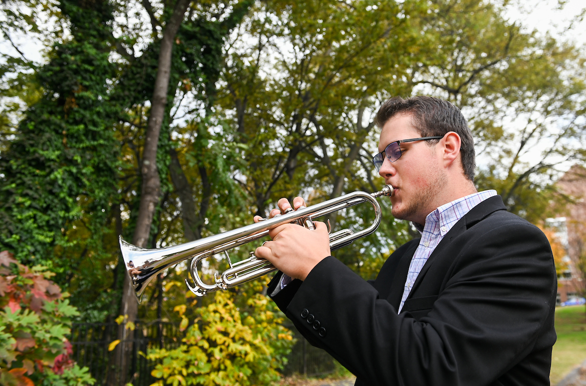 A man in a suit plays a silver trumpet outdoors.
