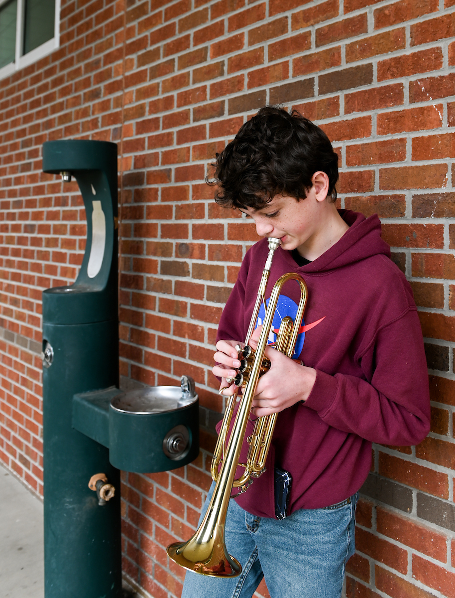 Young boy playing a trumpet and standing against a brick wall near a green drinking fountain.