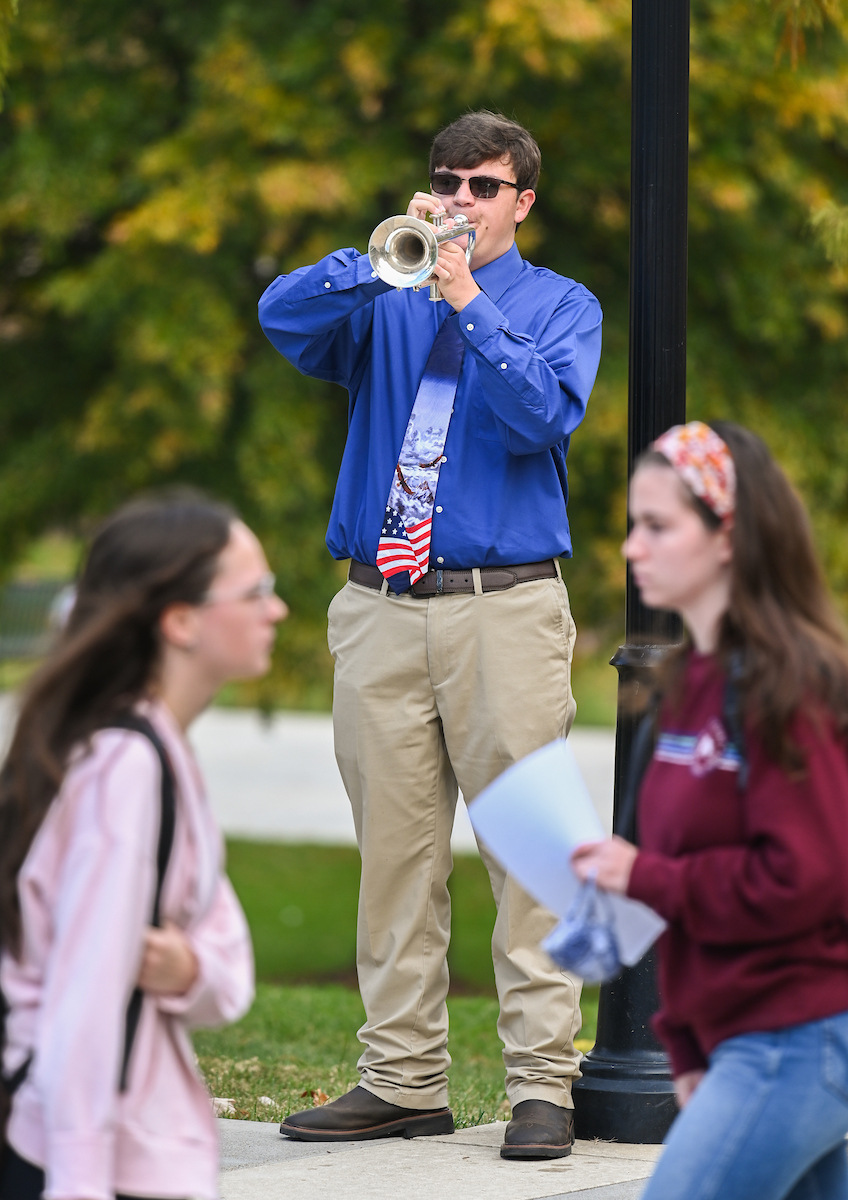 A man plays a trumpet outside, wearing a blue shirt, flag-patterned tie, and sunglasses. Two women walk by, one holding papers.