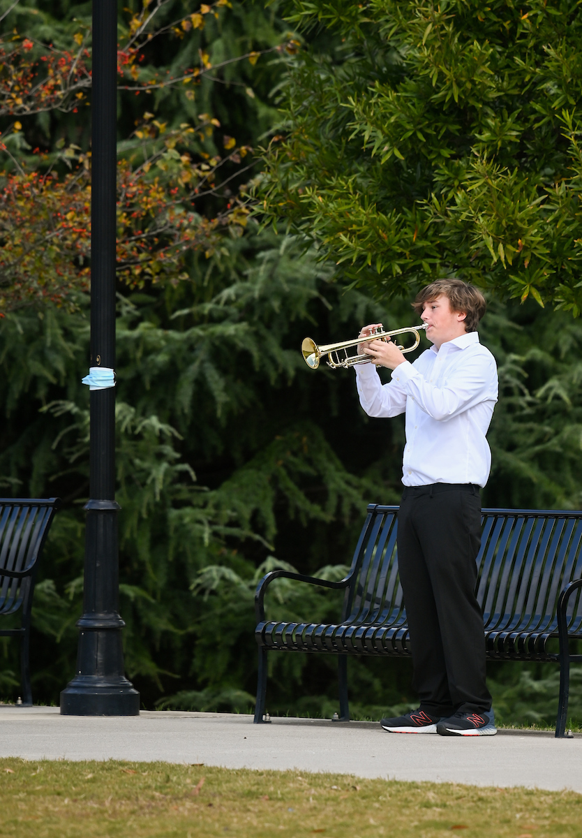 A person in a white shirt plays a trumpet outdoors. he stand s near a bench and lamppost.