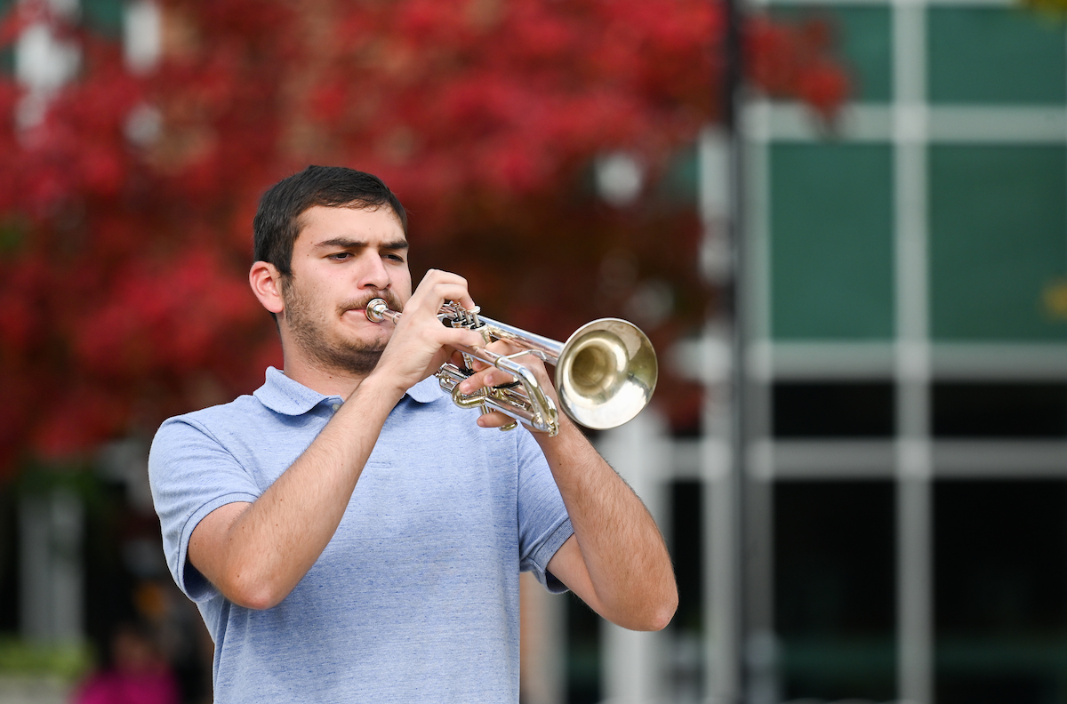 A man in a light blue polo shirt plays a silver trumpet outdoors.