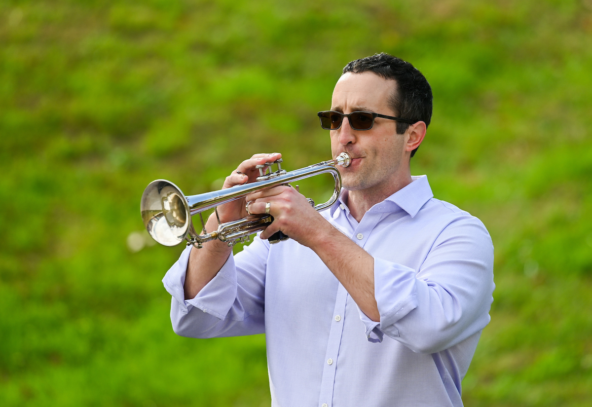 A man wearing a buttoned-up shirt and sunglasses plays a silver trumpet outdoors, in a grassy area.