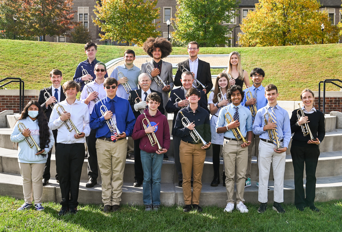 A group of people stand outdoors on steps, each holding a trumpet.