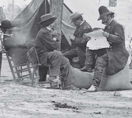 Four men in hats and coats sit around outside. One reads a newspaper while others converse.