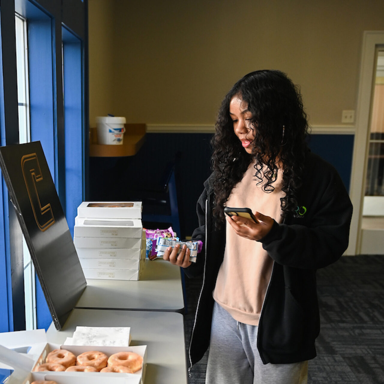 A woman with curly hair holding a phone and snacks, standing beside a table stacked with donut boxes.