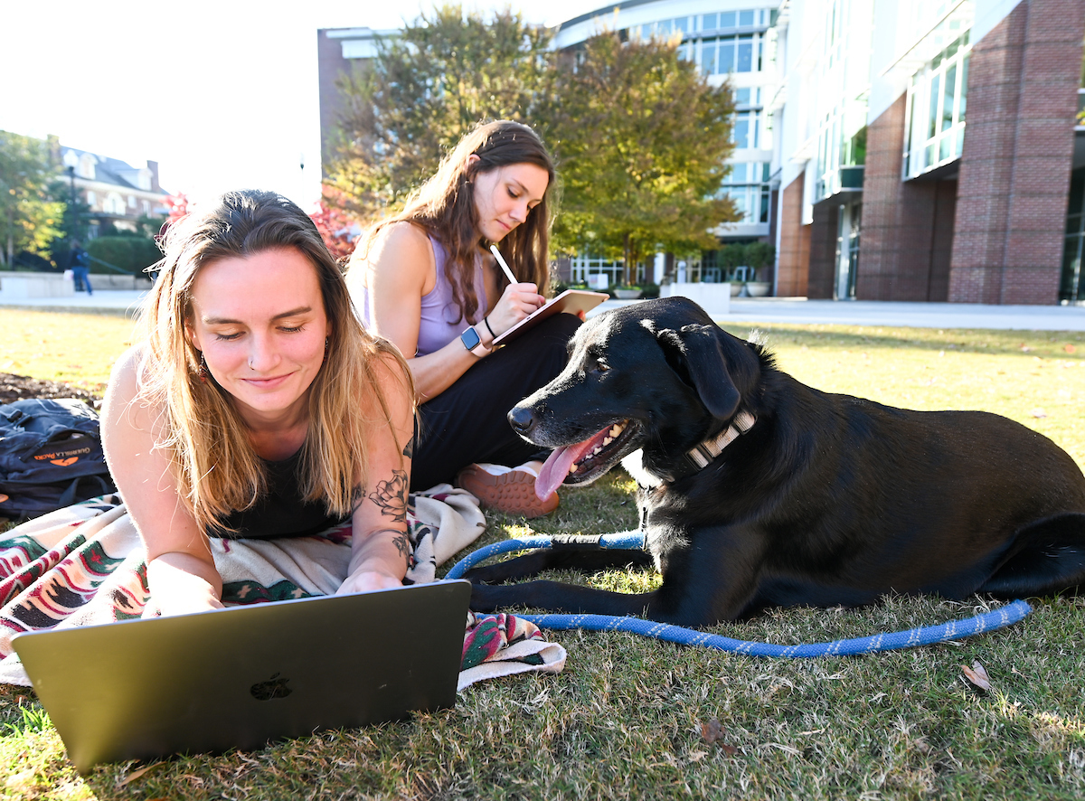 Students relax on campus, along with a friendly dog, on Chamberlain Field.