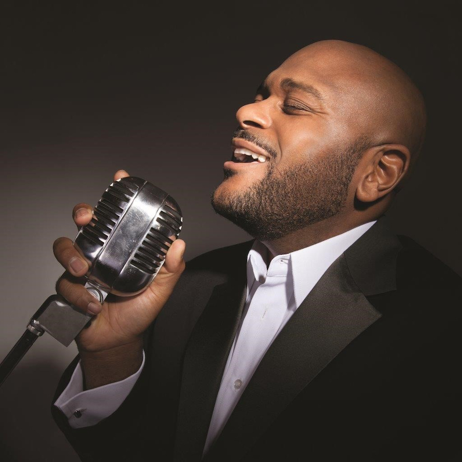 Ruben Studdard singing passionately into a vintage microphone against a dark background.