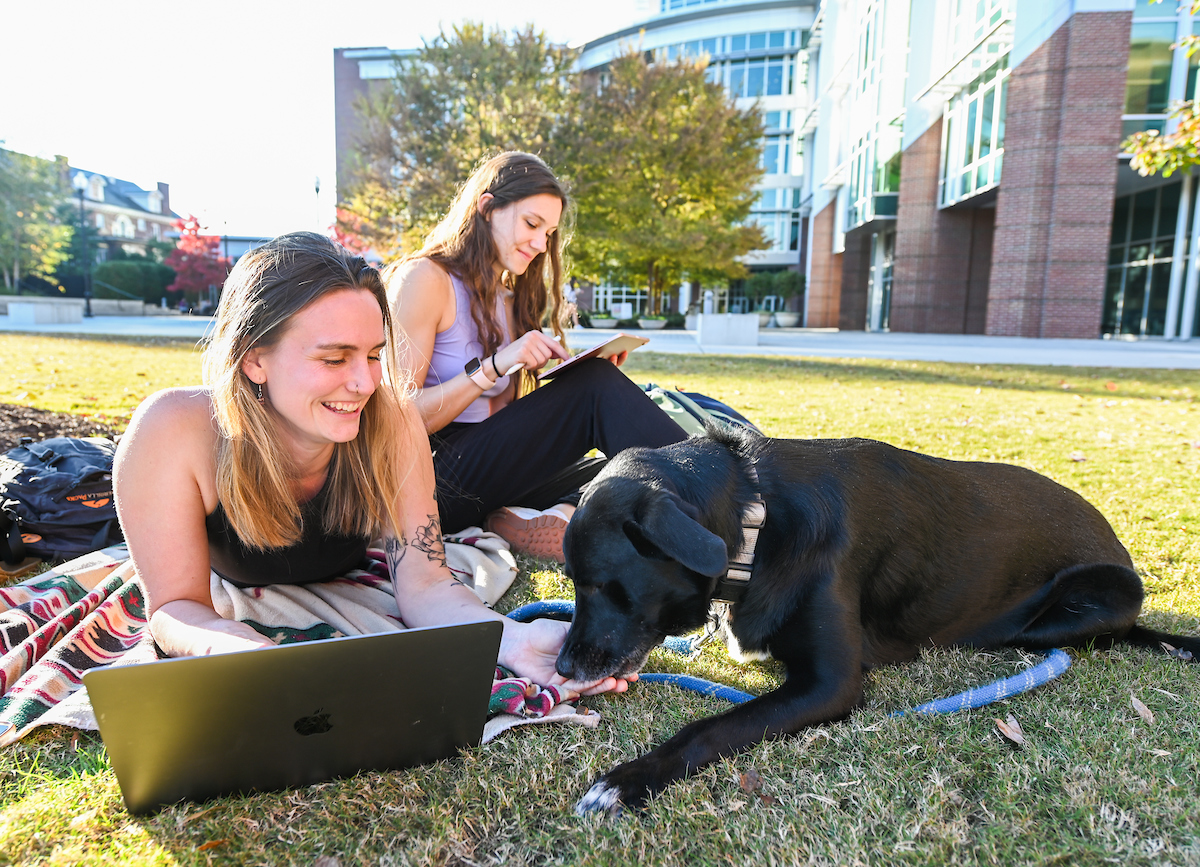 UTC students relaxing on the Chamberlain Field. A black dog sits beside them.