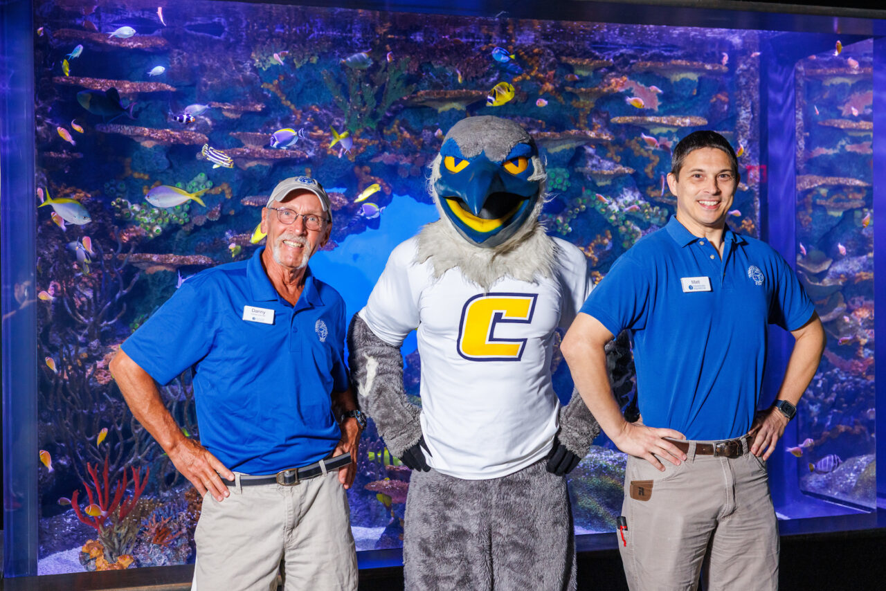 University of Tennessee at Chattanooga Alumni and Senior Aquarist Danny Alexander, left, and Curator of Fishes Matt Hamilton pose with mascot Scrappy in front of the Indo-Pacific Reef exhibit at the Tennessee Aquarium.