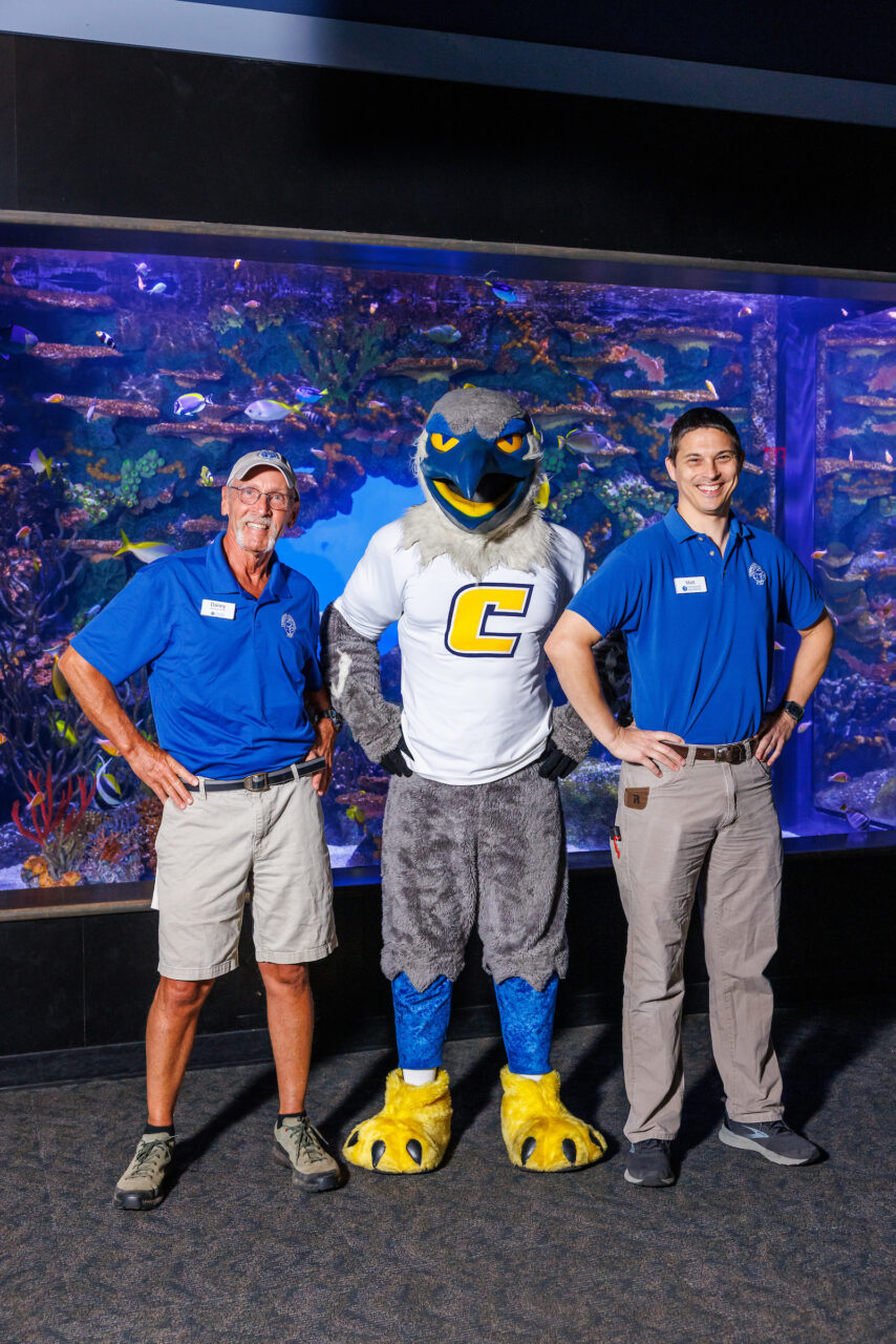 University of Tennessee at Chattanooga Alumni and Senior Aquarist Danny Alexander, left, and Curator of Fishes Matt Hamilton pose with mascot Scrappy in front of the Indo-Pacific Reef exhibit at the Tennessee Aquarium.