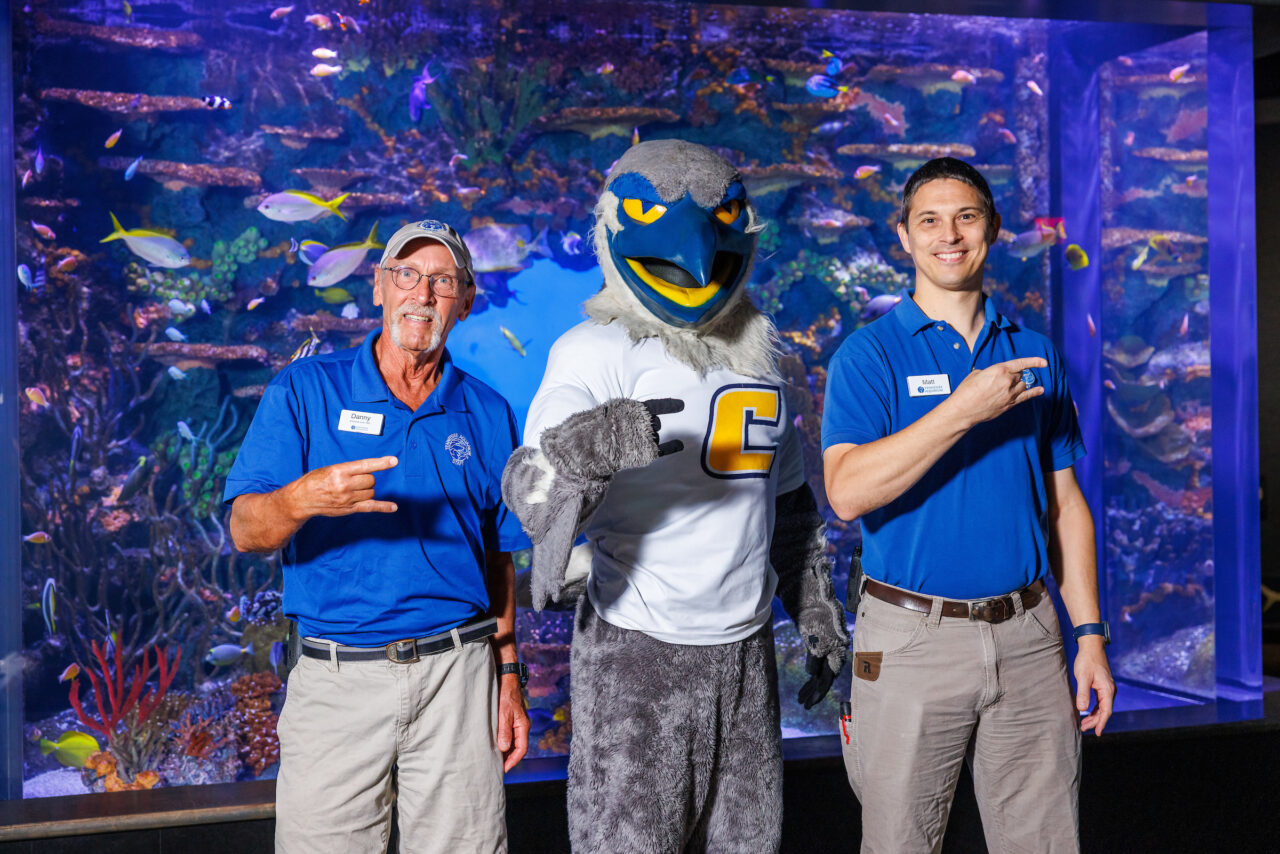 University of Tennessee at Chattanooga Alumni and Senior Aquarist Danny Alexander, left, and Curator of Fishes Matt Hamilton pose with mascot Scrappy in front of the Indo-Pacific Reef exhibit at the Tennessee Aquarium.