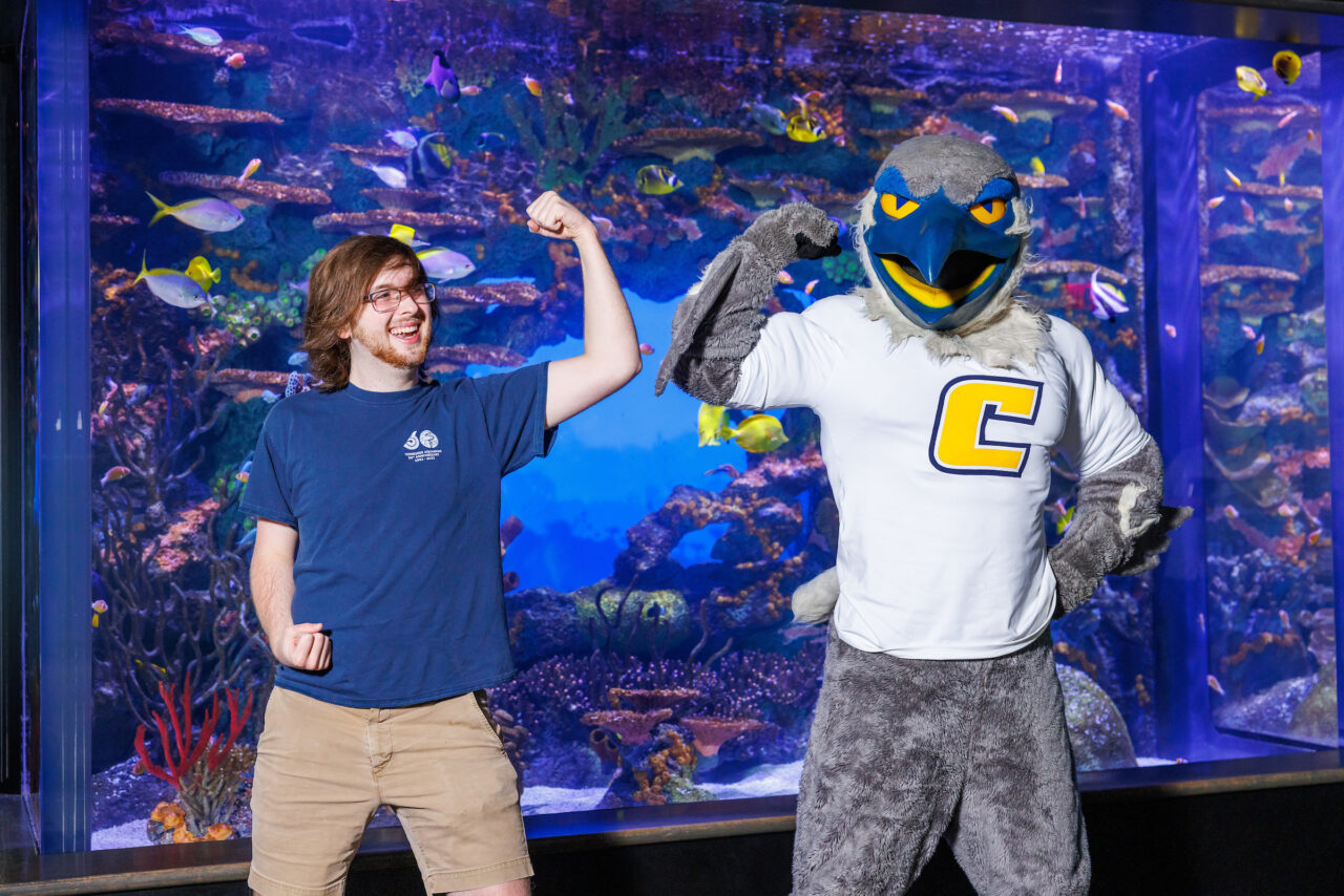 University of Tennessee at Chattanooga student and Reintroduction Assistant Seth Little poses with mascot Scrappy in front of the Indo-Pacific Reef exhibit at the Tennessee Aquarium.