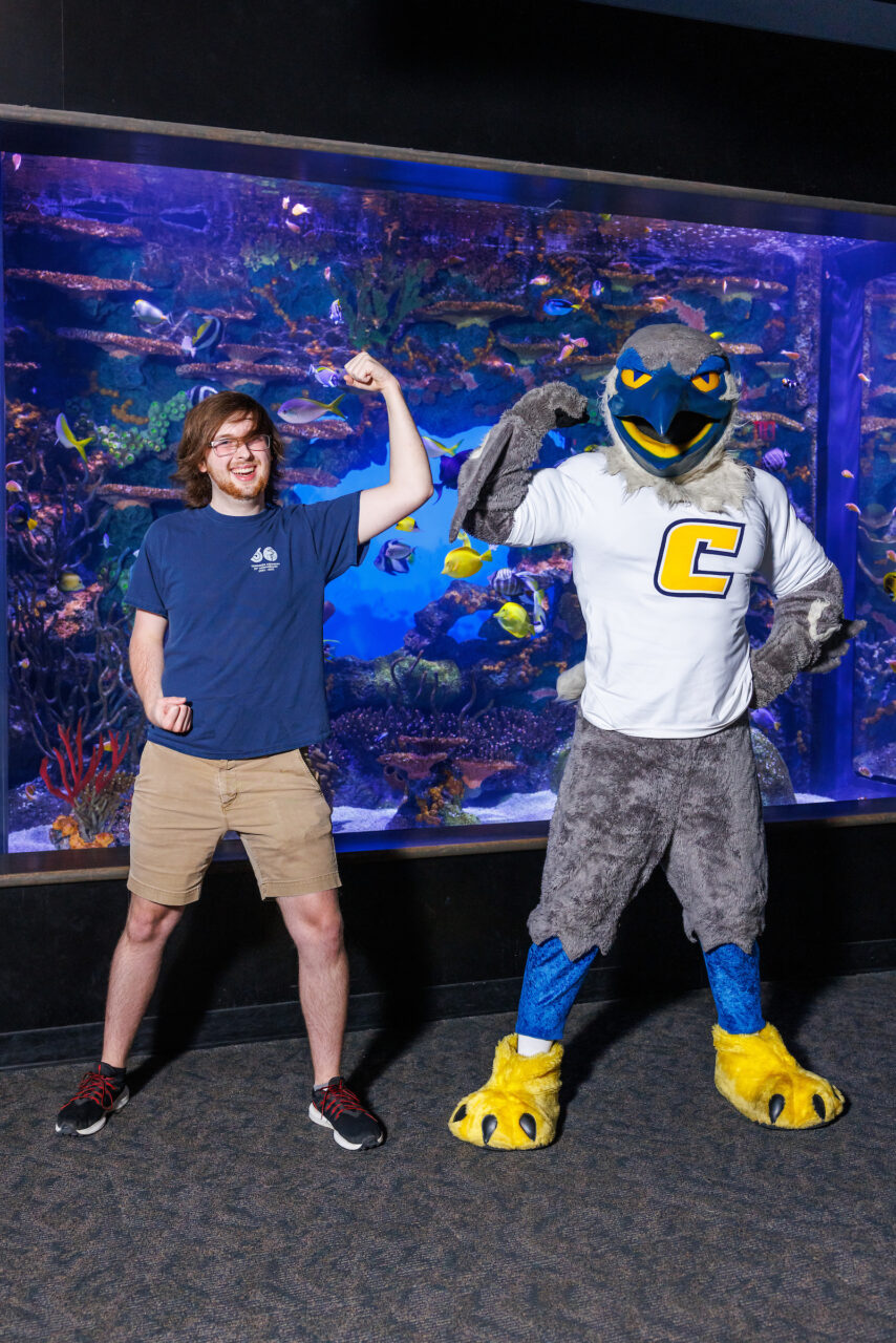 University of Tennessee at Chattanooga student and Reintroduction Assistant Seth Little poses with mascot Scrappy in front of the Indo-Pacific Reef exhibit at the Tennessee Aquarium.
