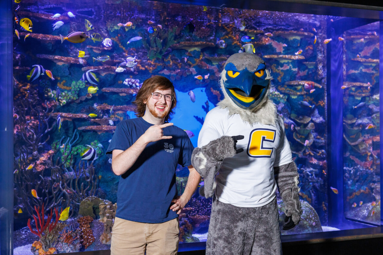 University of Tennessee at Chattanooga student and Reintroduction Assistant Seth Little poses with mascot Scrappy in front of the Indo-Pacific Reef exhibit at the Tennessee Aquarium.