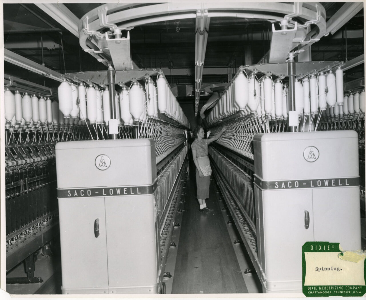 Photograph depicting an employee standing among a row of machines and skeins of yarn in a Dixie Mercerizing Company plant, Chattanooga, Tennessee. The photograph is labeled: "Spinning."