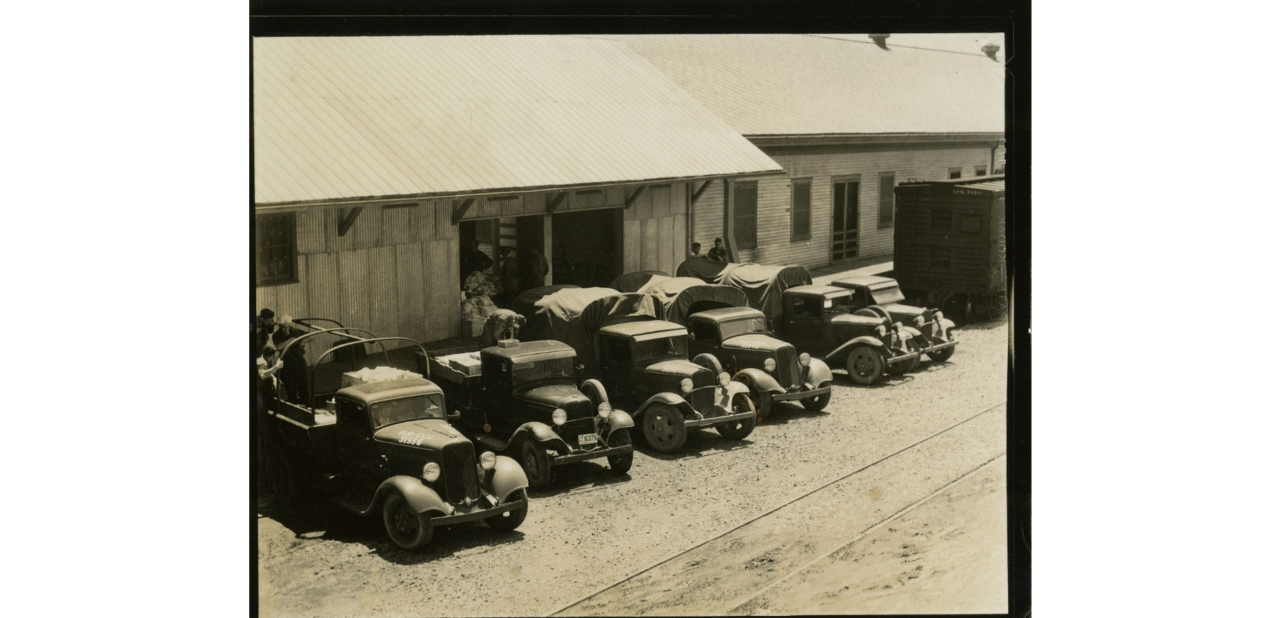 Civilian Conservation Corps photograph from the 1930s of six trucks, several workers and a railroad boxcar at a loading dock.