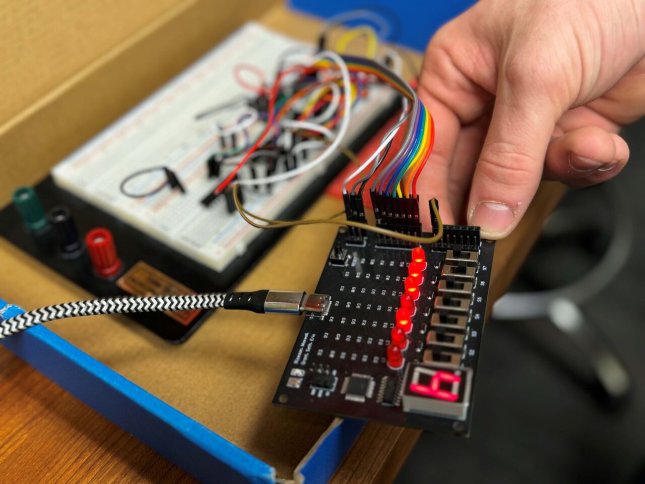 The black object in the foreground is the UTC student-developed DigiLab One logic board for use in digital logic lab courses. The red lights indicate the output states generated by a circuit, multicolored wires connect the board to circuits, and the black-and-white patterned cable enables the board to interact with the computer software to run automated testing.