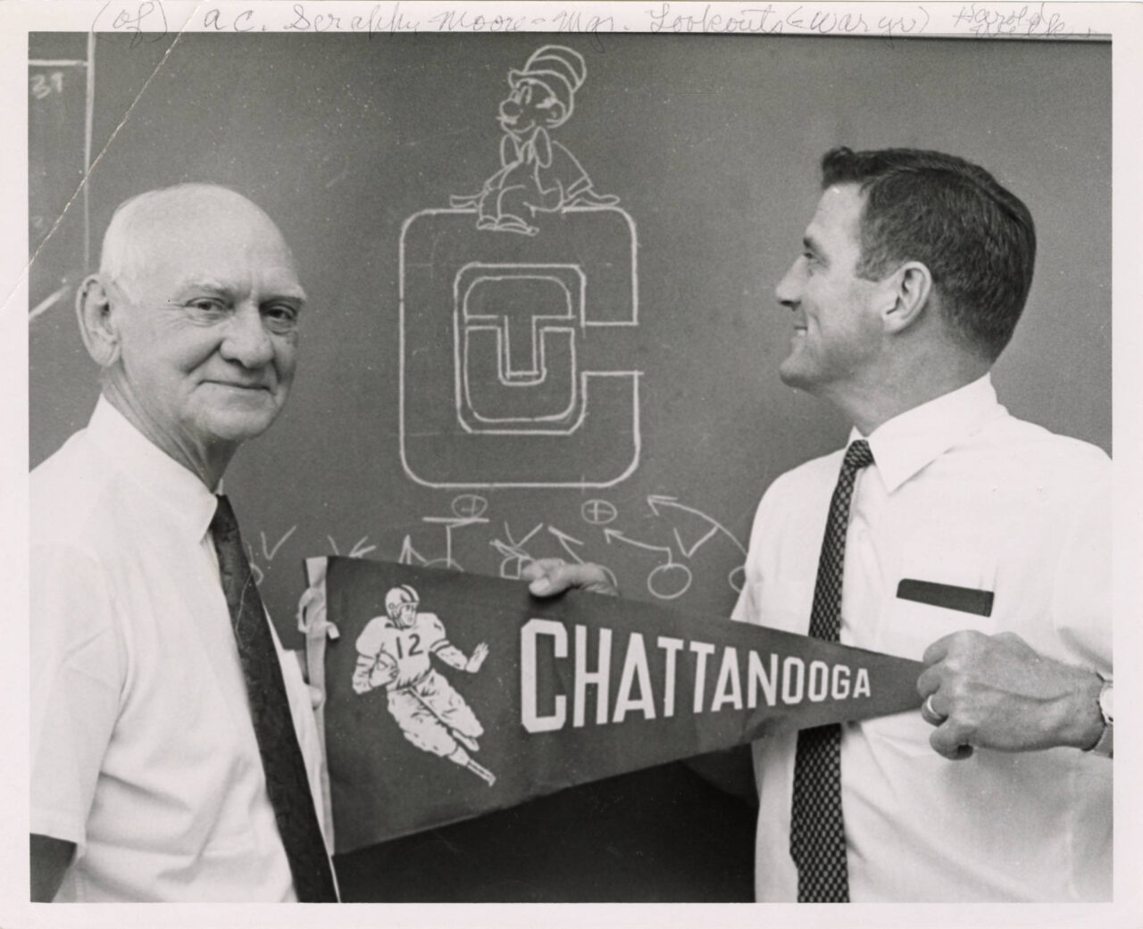 Black-and-white photograph of former University of Chattanooga football coach and Athletic Director Scrappy Moore with his successor, Harold Wilkes, holding a University of Chattanooga football pennant. Photo courtesy of University of Tennessee at Chattanooga Special Collections.