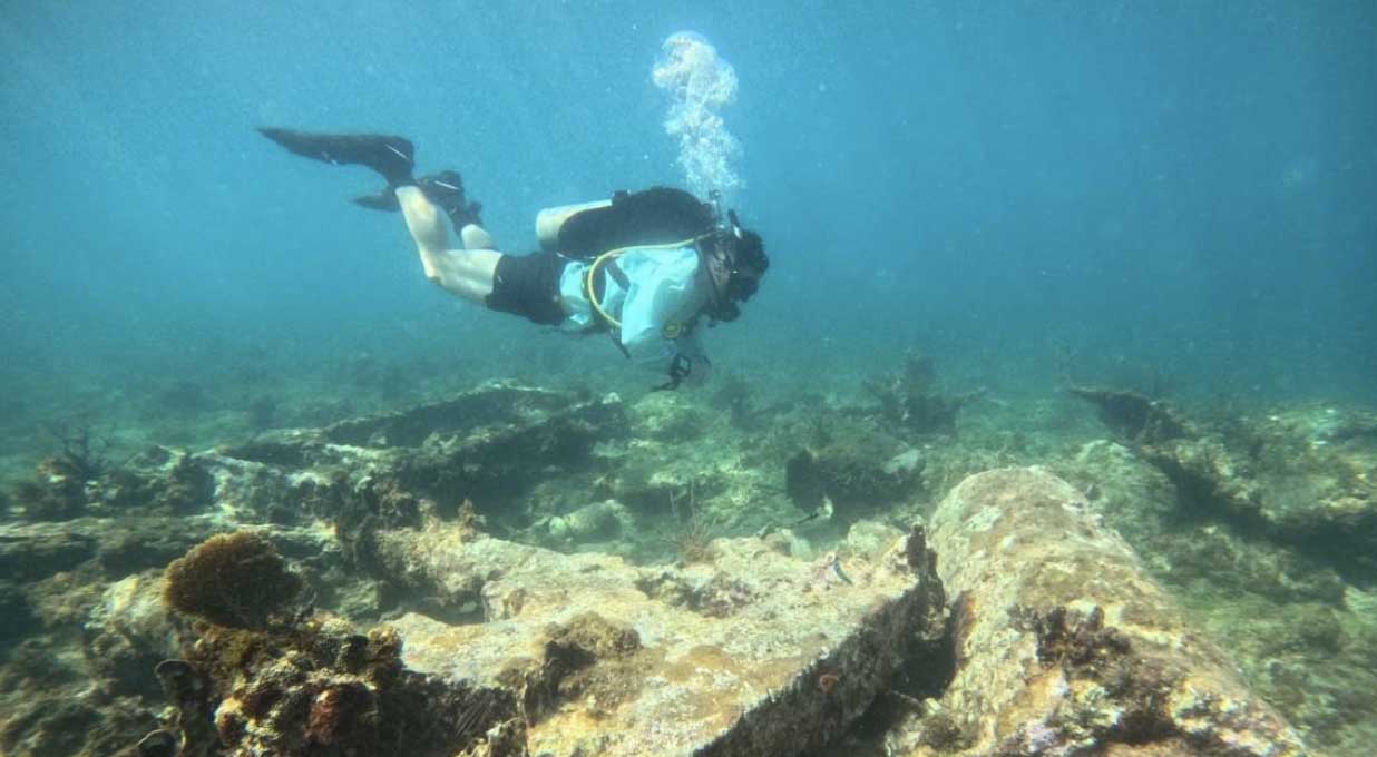 Andrew J. Van Slyke dives under the water with diving gear. Traces of the Sugar Mill Wreck are on the sea floor.
