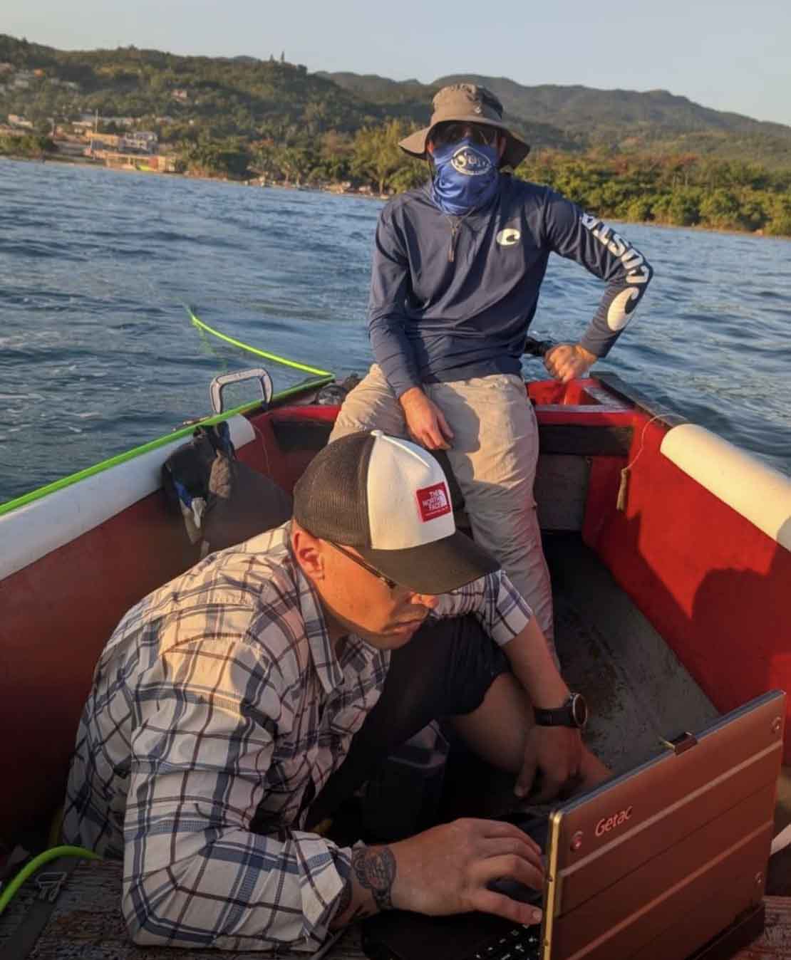 Two men sit in a red boat on the water. Morgan operates the boat and Andrew is looking at a laptop.