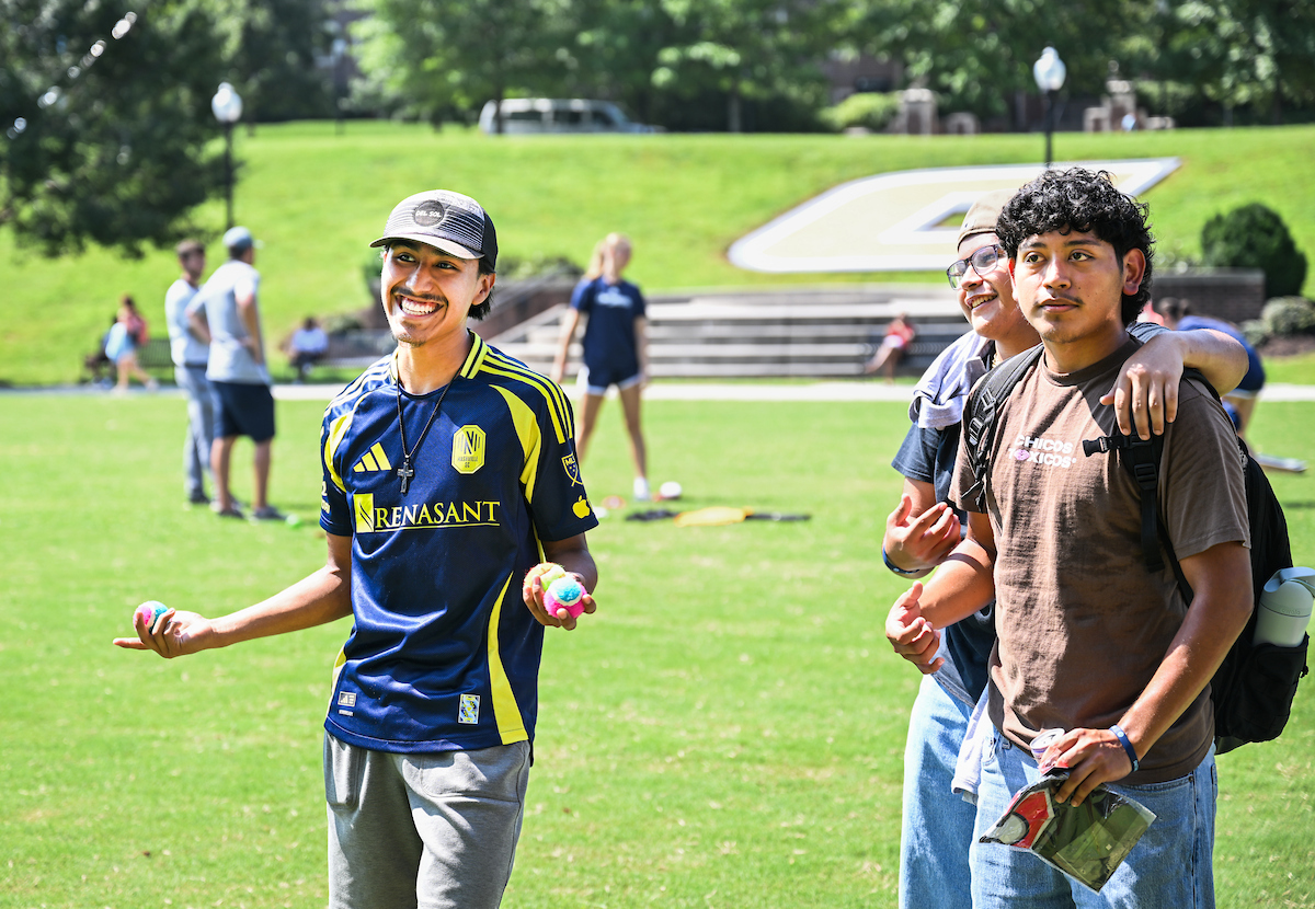 Three students smile and pose together on Chamberlain Field during an outdoor campus event. One holds small colorful balls, while the others stand with backpacks and water bottles.