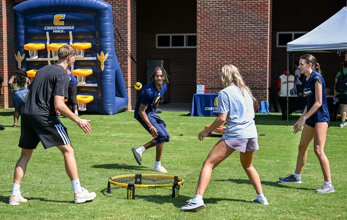 Four students play Spikeball on a grassy field during a campus event. A large inflatable basketball game with UTC branding is visible in the background.