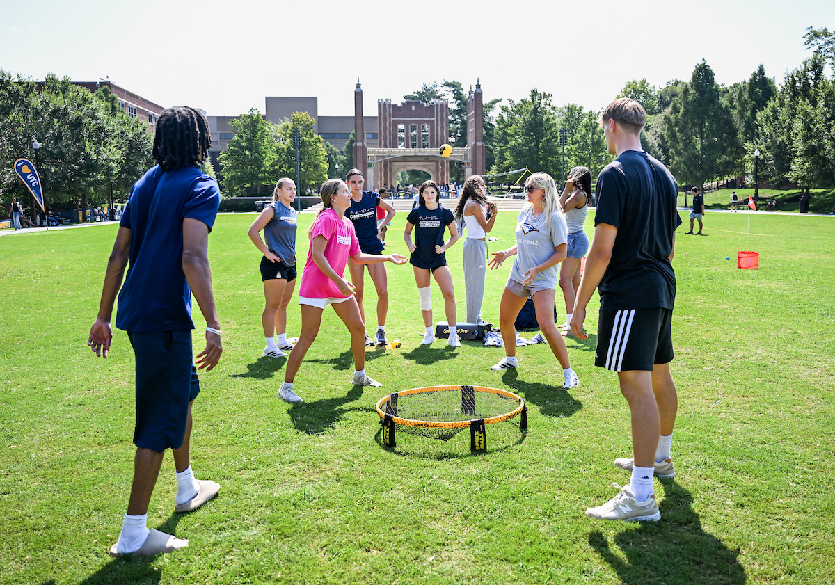 A group of students gathers around a Spikeball net on Chamberlain Field, with some actively playing and others watching. UTC’s entrance arch is visible in the background.