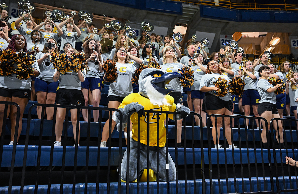 Scrappy the mascot against the rail with the band performing behind him in the stands
