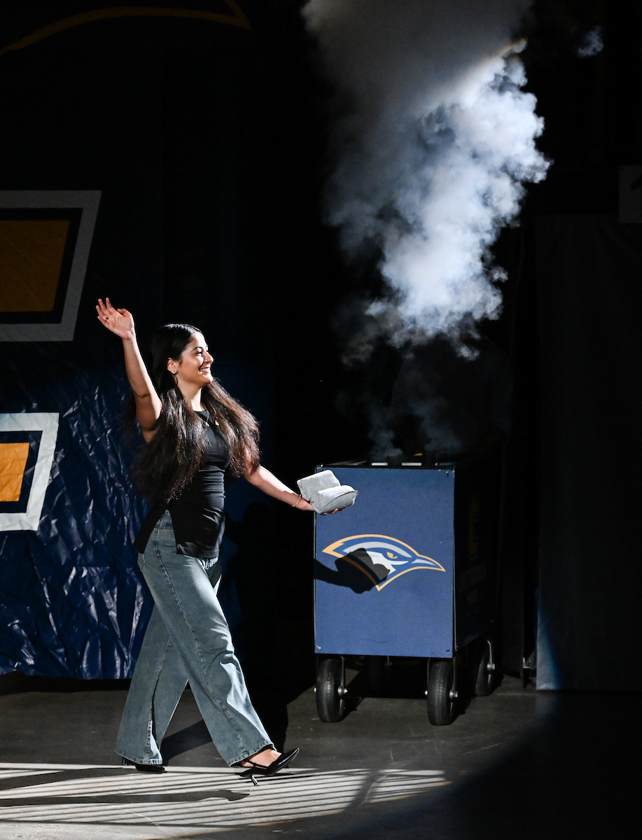 Female student walking out of "tunnel" with a Power C on it. She is waving to the crowd and holding t-shirts to throw. Fog billows in the background.