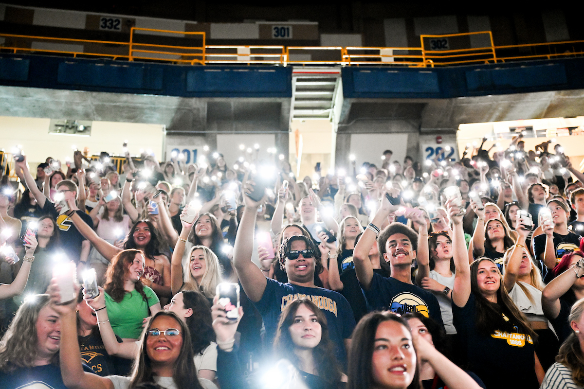 A crowd of students hold up their phone flashlights in McKenzie Arena
