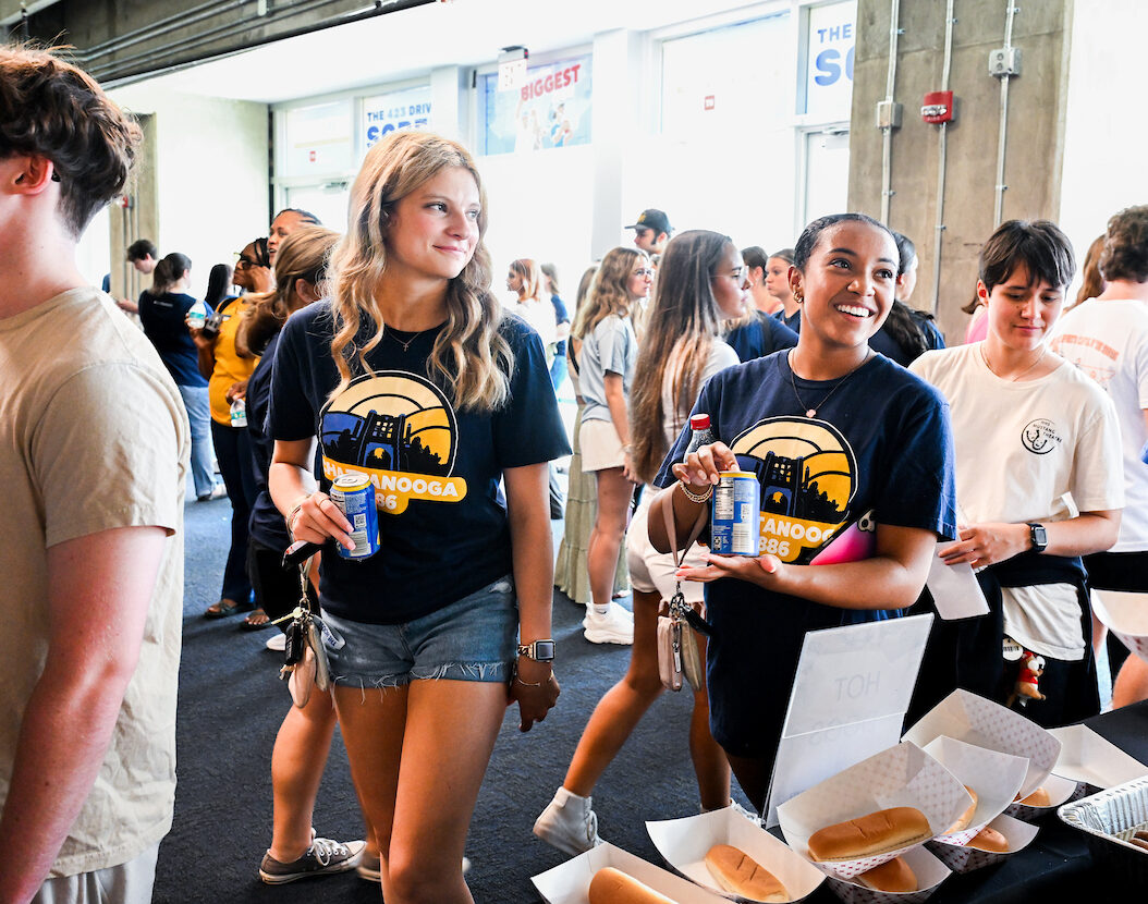 Two students smile as they pick up food from a table with hotdogs. A crowd of other students stands behind them in McKenzie Arena