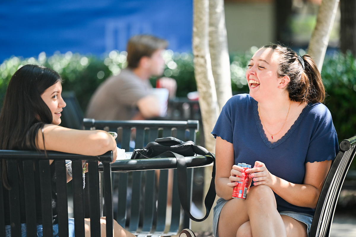 Two students sit outside at a table, talking and laughing.