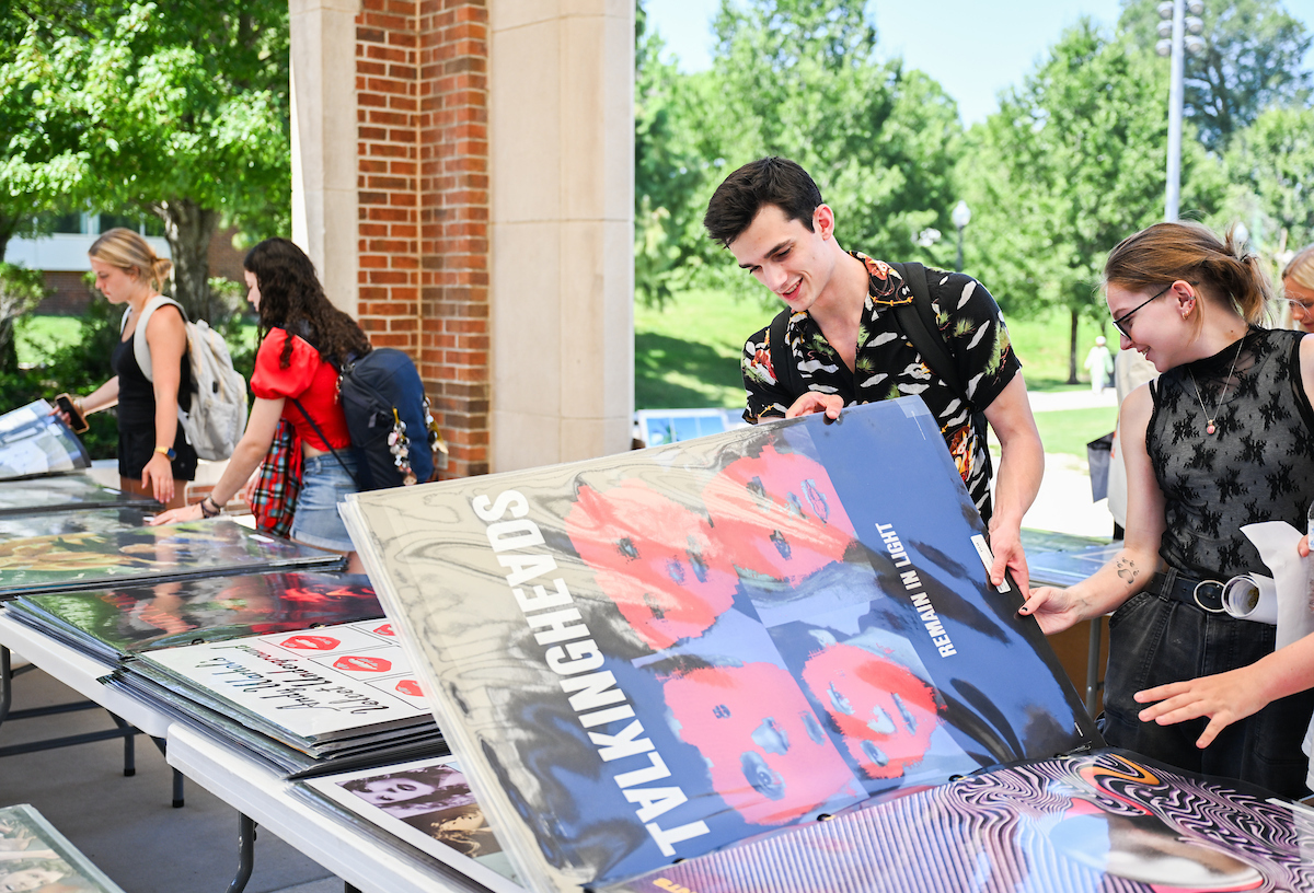 Students look through posters displayed on tables outside; one student holds up a large Talking Heads poster.