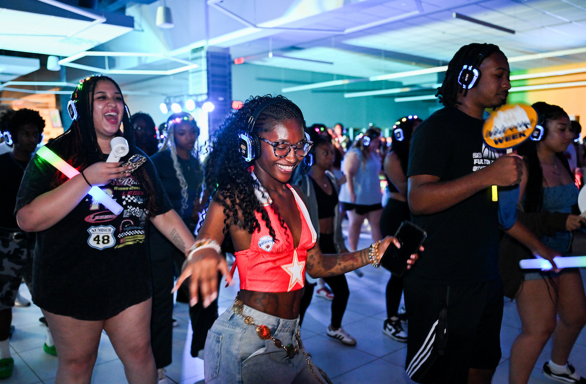 A group of students wearing headphones dance together, smiling and raising light-up sticks in the air.
