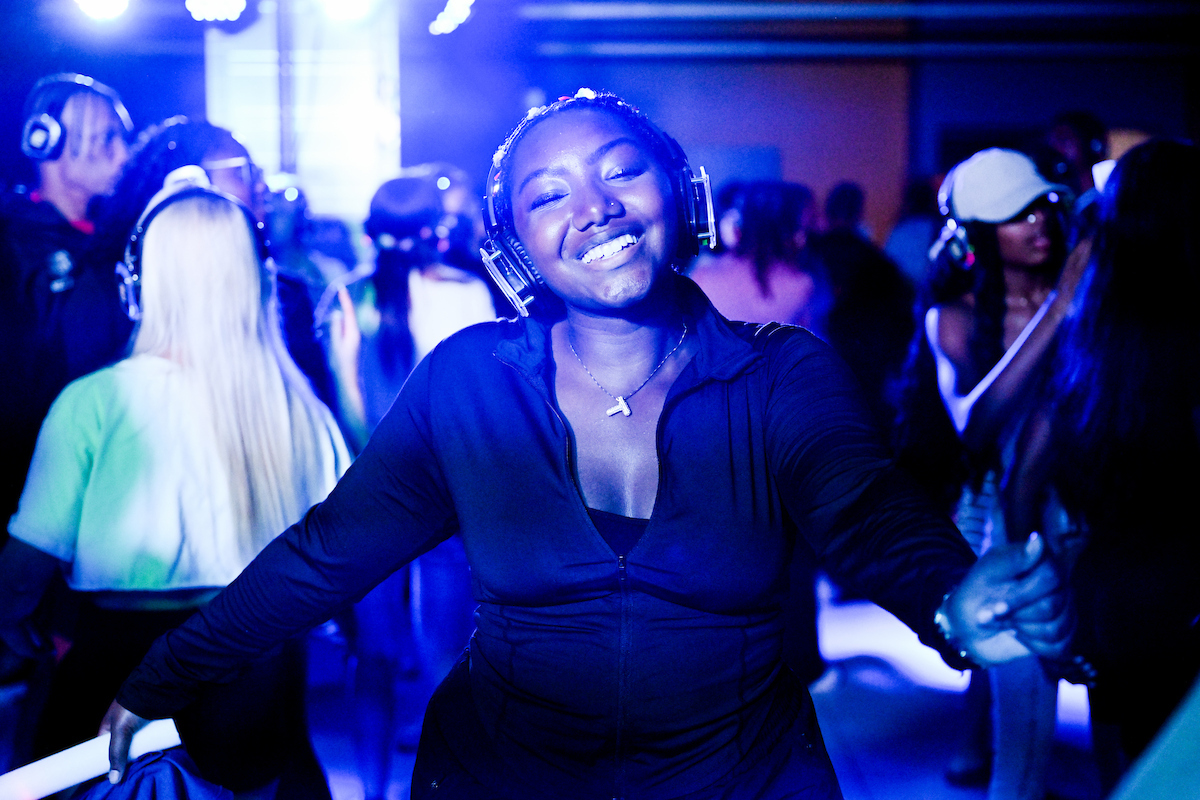 A student wearing headphones smiles directly at the camera while dancing under blue lighting.