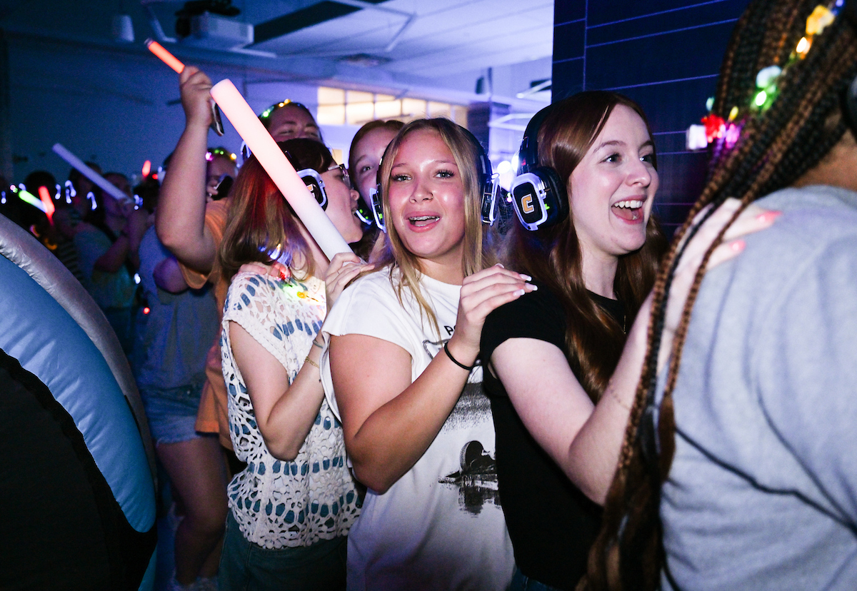 Students wearing glowing headphones dance in a line with hands on each other’s shoulders, holding light-up sticks.