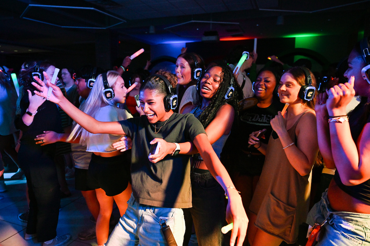 A student wearing headphones smiles while holding a light-up stick in the middle of a crowded dance floor.