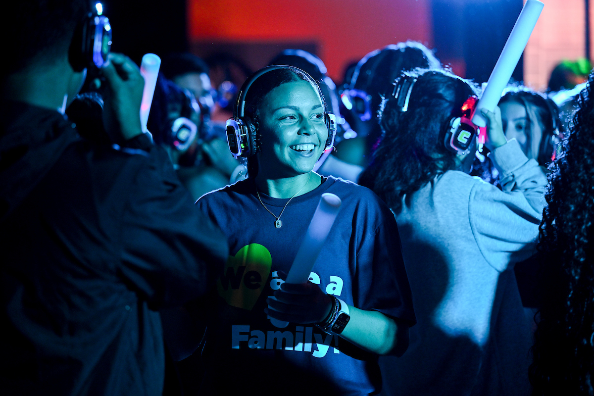 Students dance together in a crowded room, wearing headphones and holding glowing accessories.