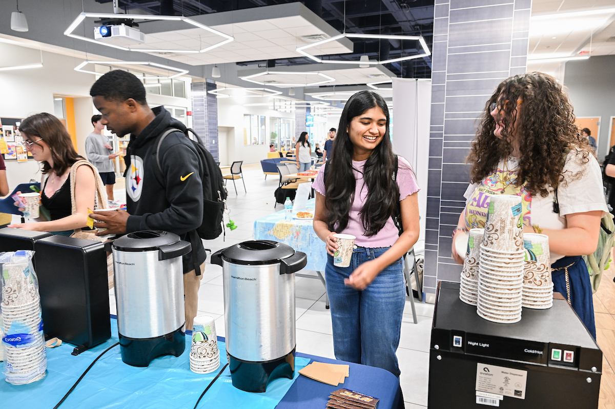 Students gather around a table with hot drink dispensers, smiling while holding cups.